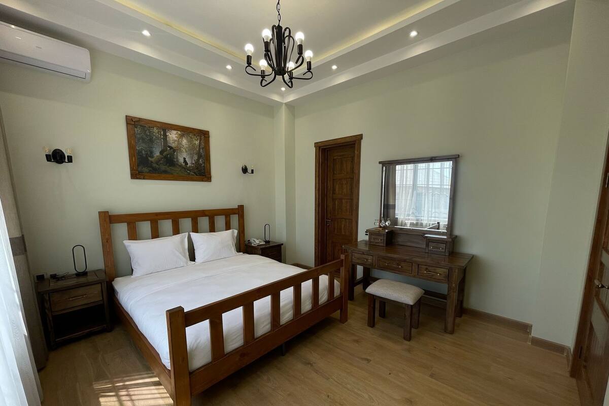 Quarto com cama de madeira, escrivaninha com espelho, quadro na parede e lustre.