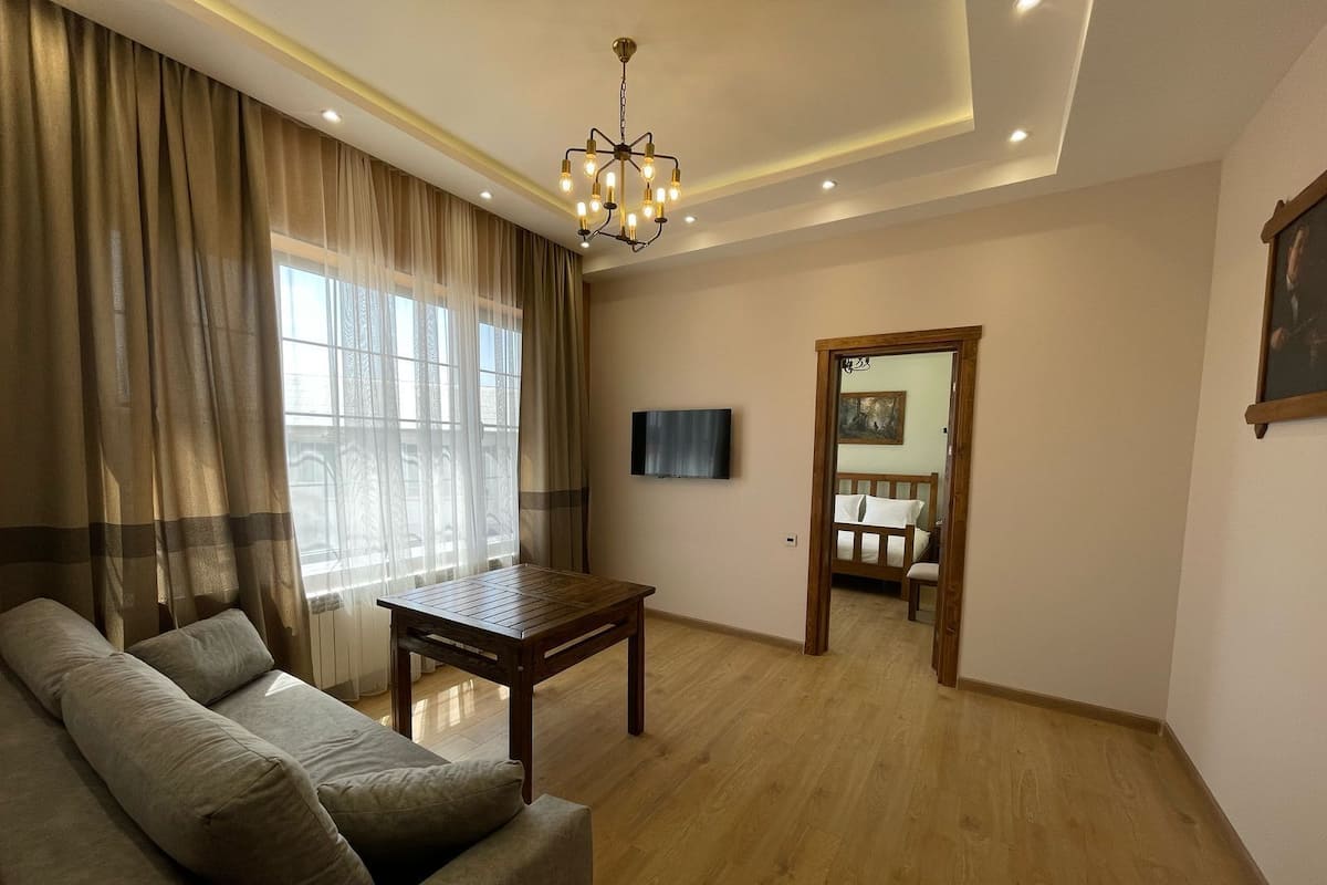 Uma sala de estar com um sofá, uma mesa de centro de madeira, uma TV fixada na parede e uma porta que dá para outro cômodo.