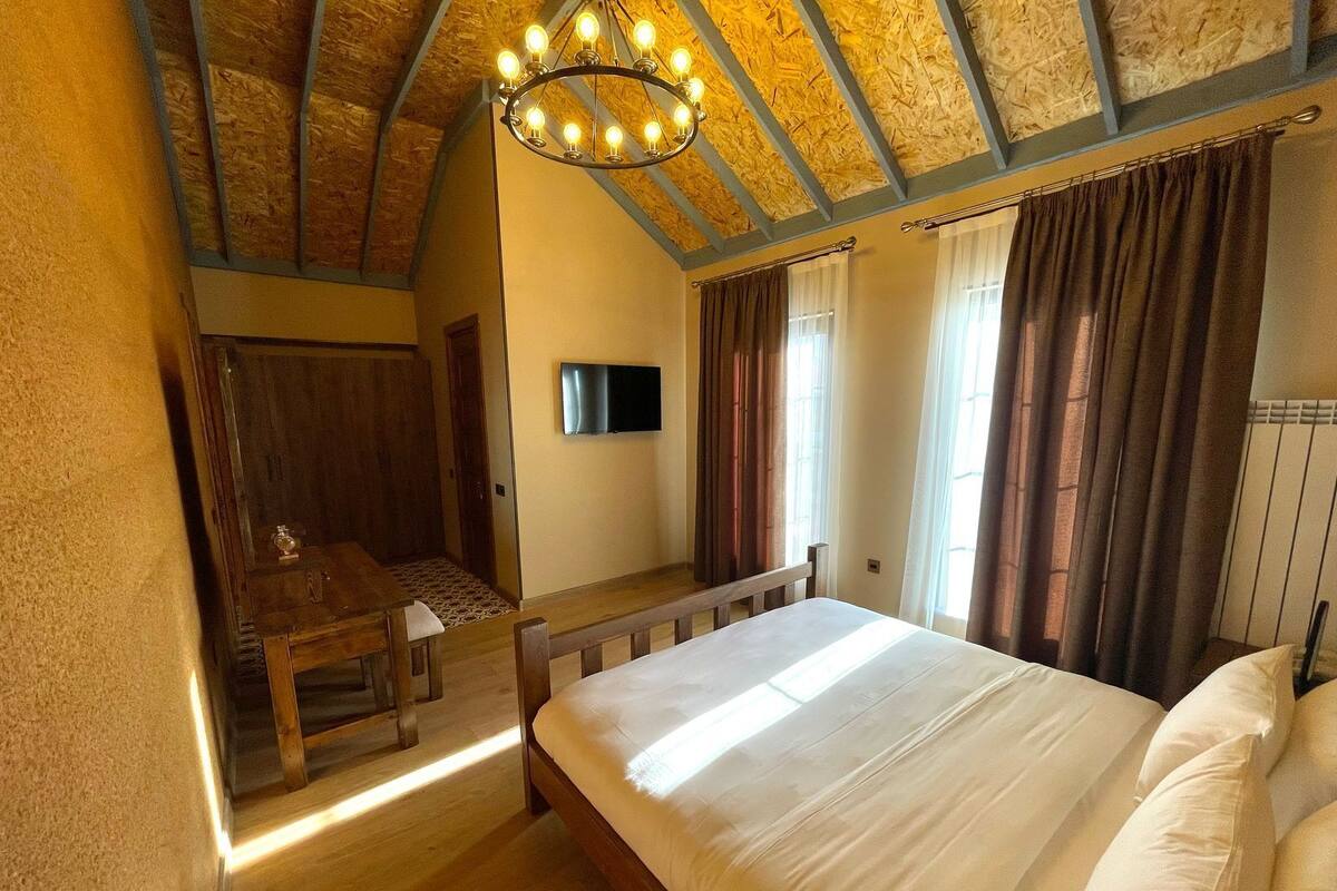 Quarto com teto de madeira, cama com roupa de cama branca, escrivaninha de madeira e televisão na parede.