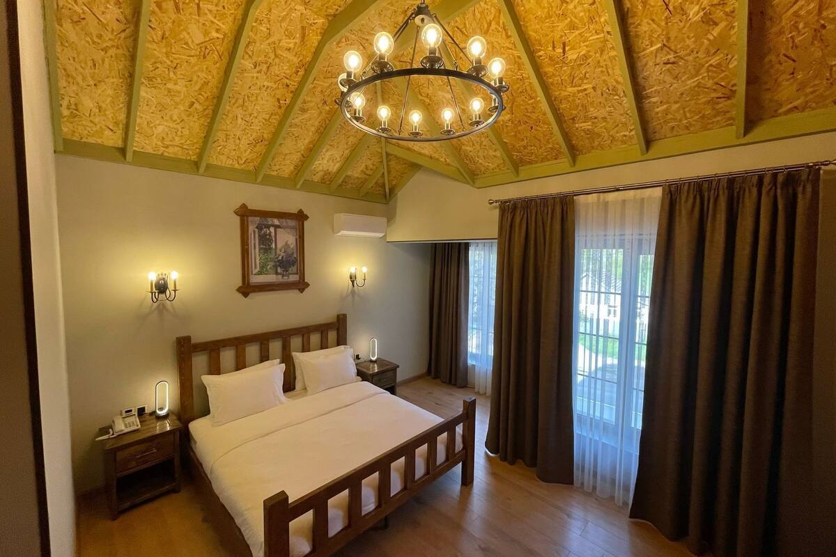 Quarto com cama de madeira, um candelabro, mesa de cabeceira, telefone e janela com cortinas.