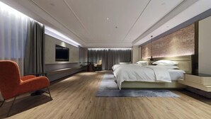 A modern hotel room with a large bed, a TV mounted on the wall, a seating area with a chair, and a wooden floor.