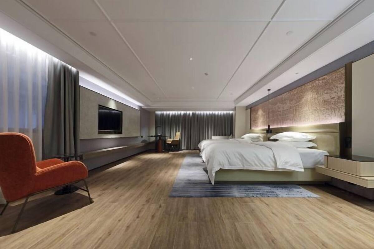 A modern hotel room with a large bed, a TV mounted on the wall, a seating area with a chair, and a wooden floor.