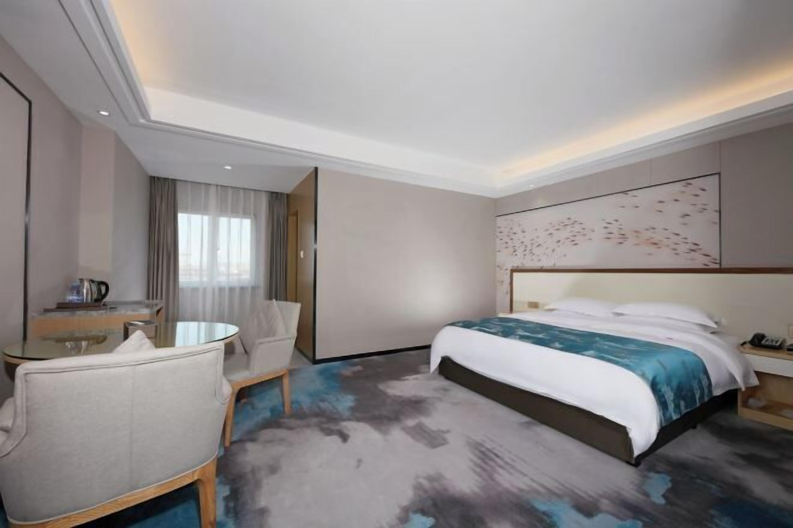A hotel room with a bed, a desk with a chair, a small table, and a large window with curtains.