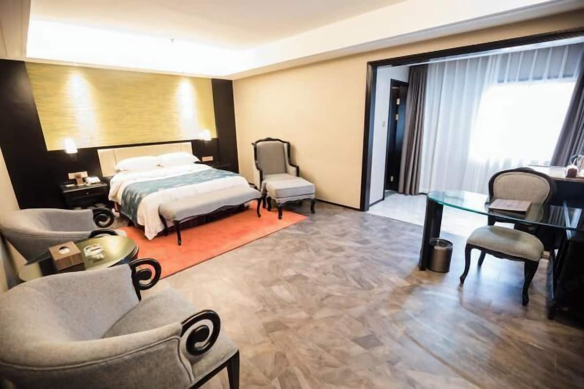 A hotel room with a bed, two armchairs, a small table, a desk, and a glass partition.