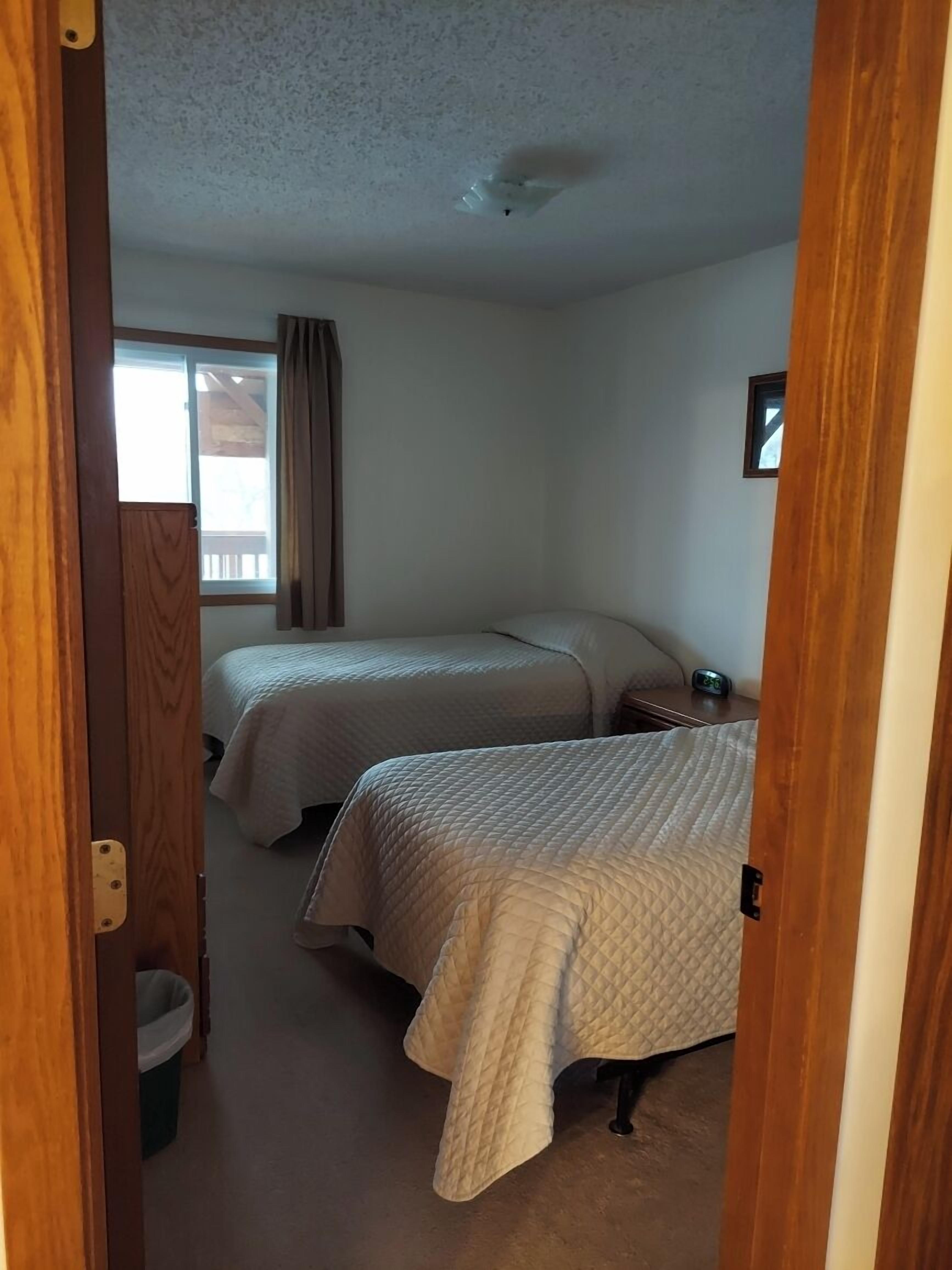 Quarto com duas camas, janela com cortinas, ventilador de teto e uma pequena mesa de cabeceira com relógio.