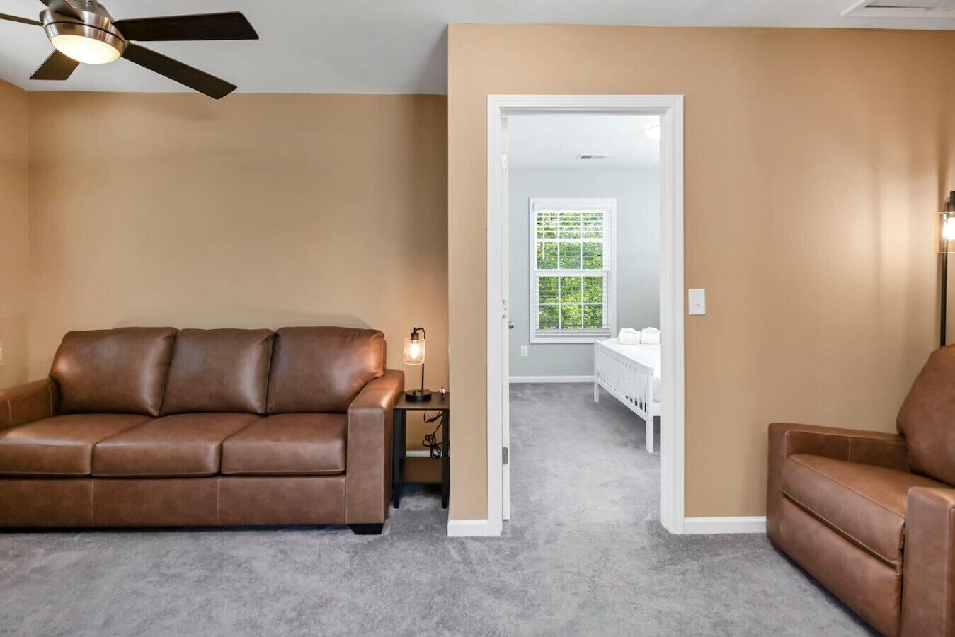 A room with a brown leather sofa, a small table with a lamp, a white crib, and a ceiling fan.