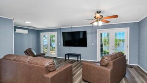 A living room with two brown leather sofas, a flat-screen TV, a ceiling fan, and a view of a lake through glass doors. - Spacious Split-Level Retreat | Sleeps 11 (Saluda County)