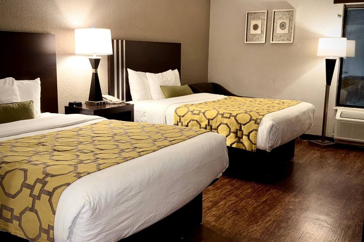 A hotel room with two beds, a nightstand, a lamp, and a window.
