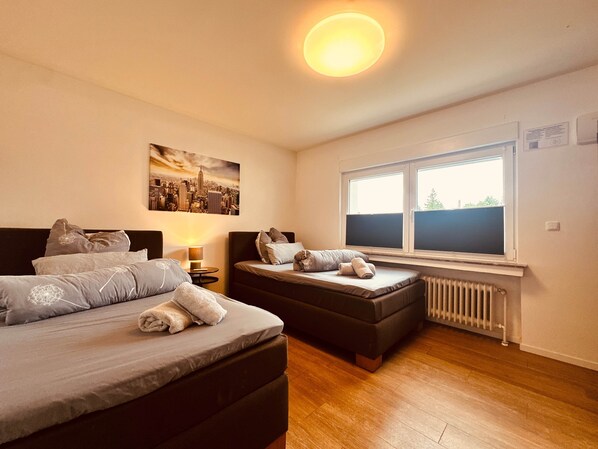 A bedroom with two beds, a cityscape painting, a window with blinds, and a radiator.