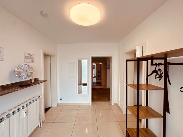 A hallway with a white wall, a radiator, a shelf with decorative items, and a wooden shelf unit with hanging items.