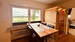 Una stanza con un tavolo in legno, sedie, un termosifone, una finestra vista su un campo e un armadietto in legno con una porta.