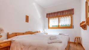 A bedroom with a wooden bed, a nightstand, a lamp, a window with a lace valance, and a small stool. - Appartamento Cordevole (Rocca Pietore)