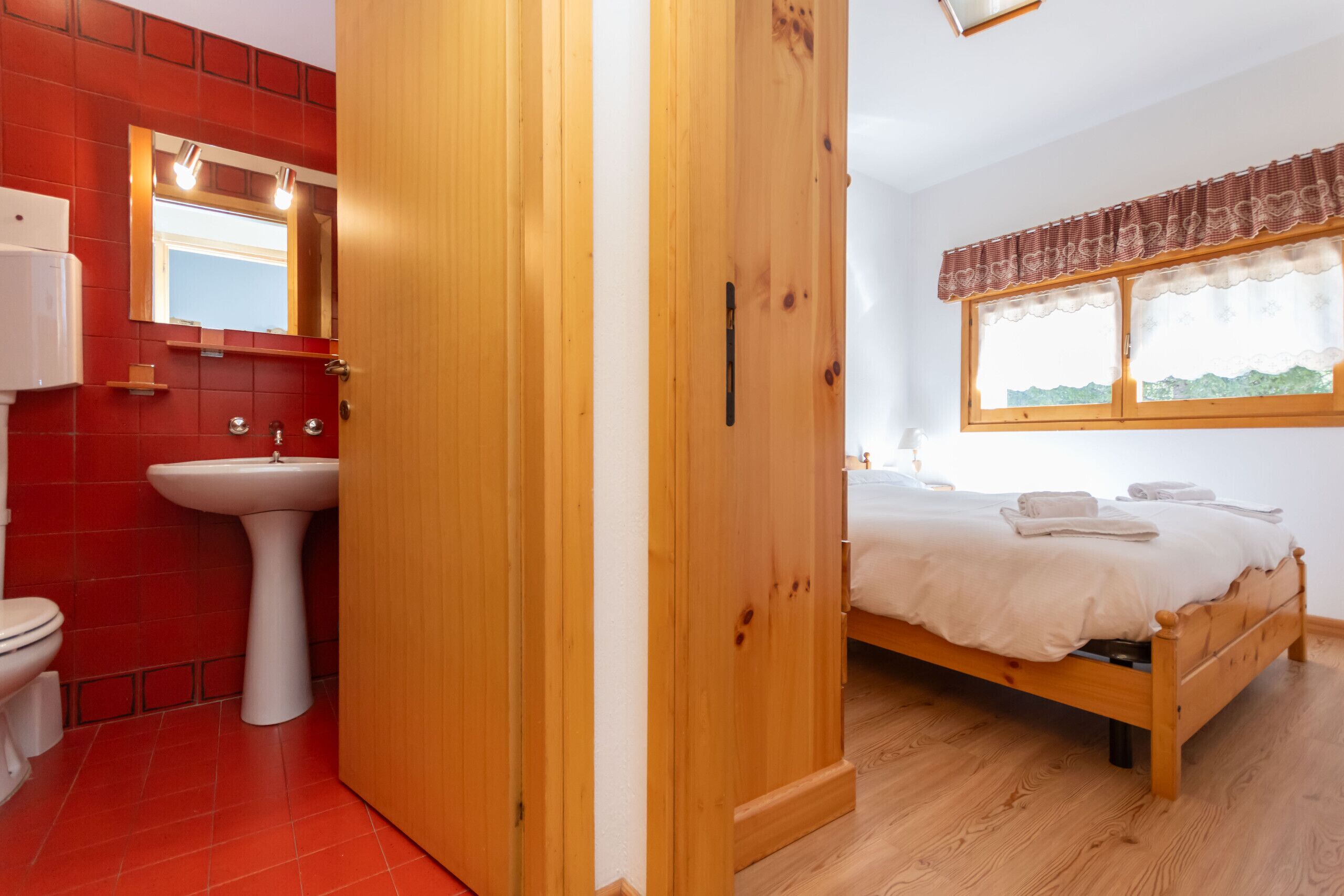 A bedroom with a wooden bed, a window with a valance, and a bathroom with a red tiled wall and a white sink.
