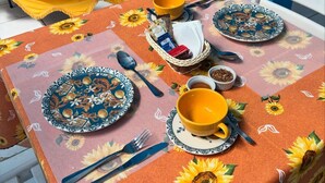 Une table dressée avec des assiettes, des bols, des couverts et un panier de collations, recouverte d’une nappe à motifs de tournesols.
