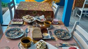Une table à manger dressée avec de la nourriture et des boissons, entourée de chaises, dans une pièce aux murs bleus et où se trouve un hamac.