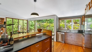 Una cocina moderna con armarios de madera, encimera negra y lavavajillas integrado. Amplias ventanas ofrecen vistas al exterior con vegetación.