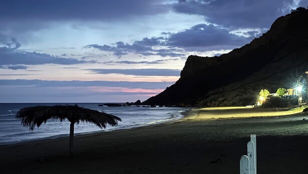 Una spiaggia con una palma, una montagna sullo sfondo e un edificio illuminato a destra.