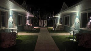 Nattebillede af et hotel med to gæsteværelser, hver med en lille patio, et bord og stole, en stenplanter og en sti til indgangen.