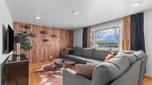 A modern living room with a grey sofa, a glass coffee table, and a wooden accent wall. - LAMBEAUView|7 MinWalk|Sleeps10 (Green Bay)