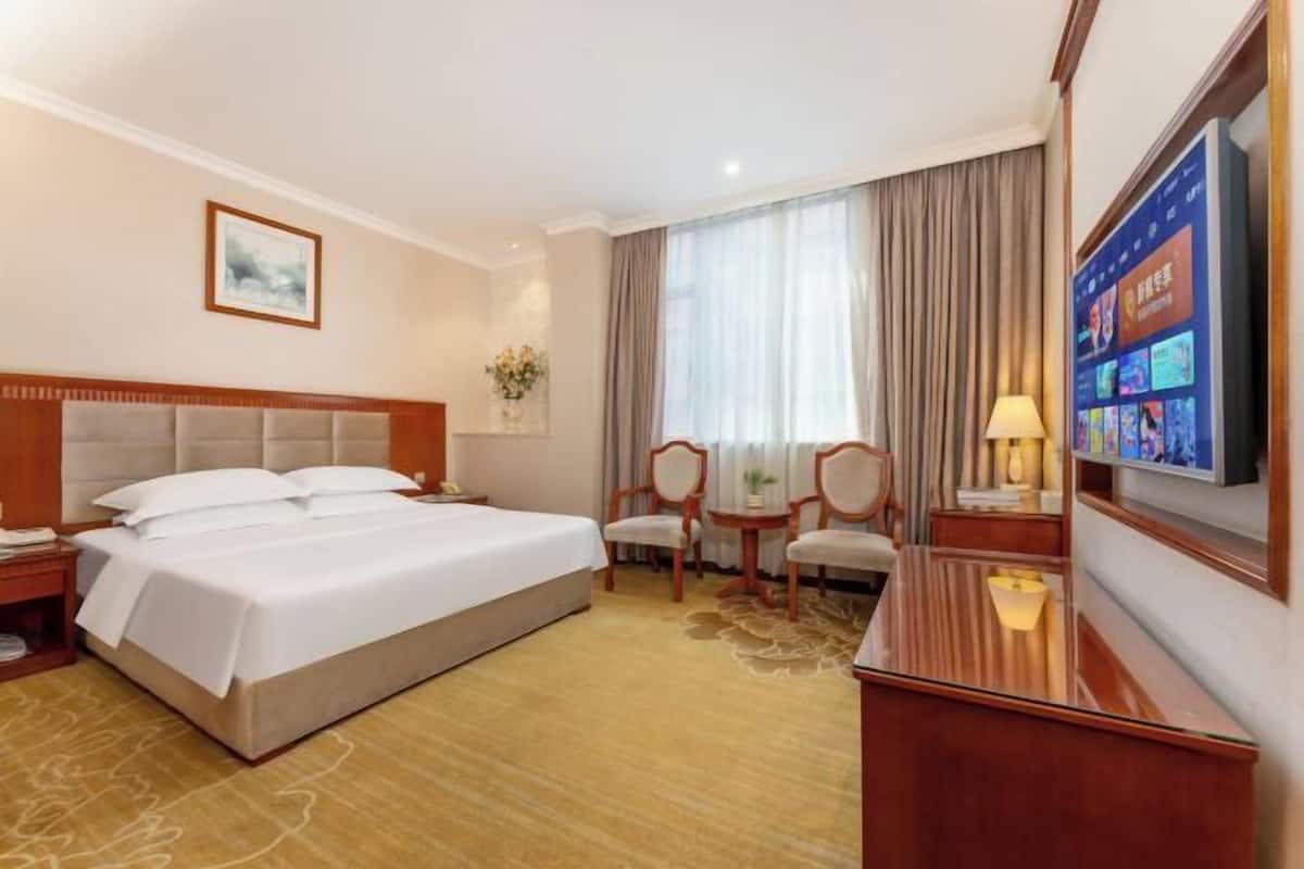 A hotel room with a large bed, a desk, a chair, a TV, and a window with curtains.