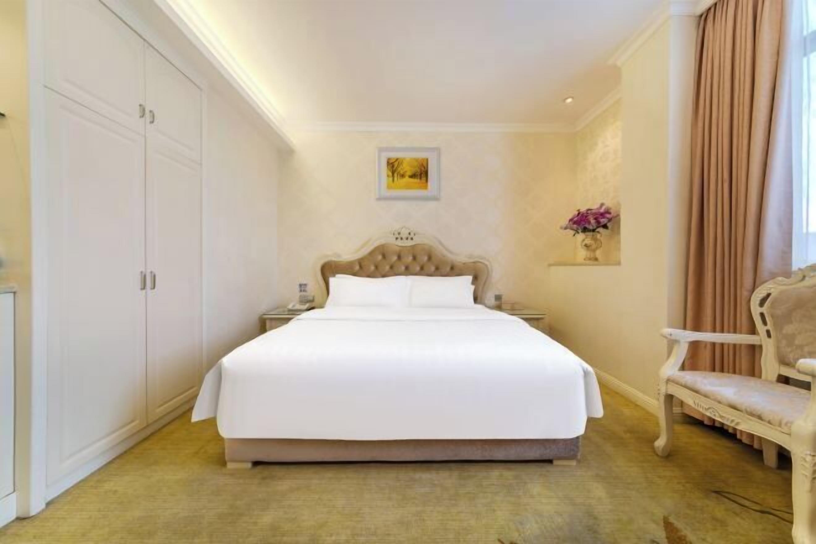 Ein Hotelzimmer mit einem großen Bett, einem Sessel, einem Nachttisch, einem Wandbild und einem Vorhang.