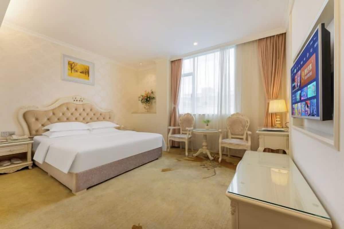 A hotel room with a large bed, a desk, a chair, a TV, and a window with curtains.