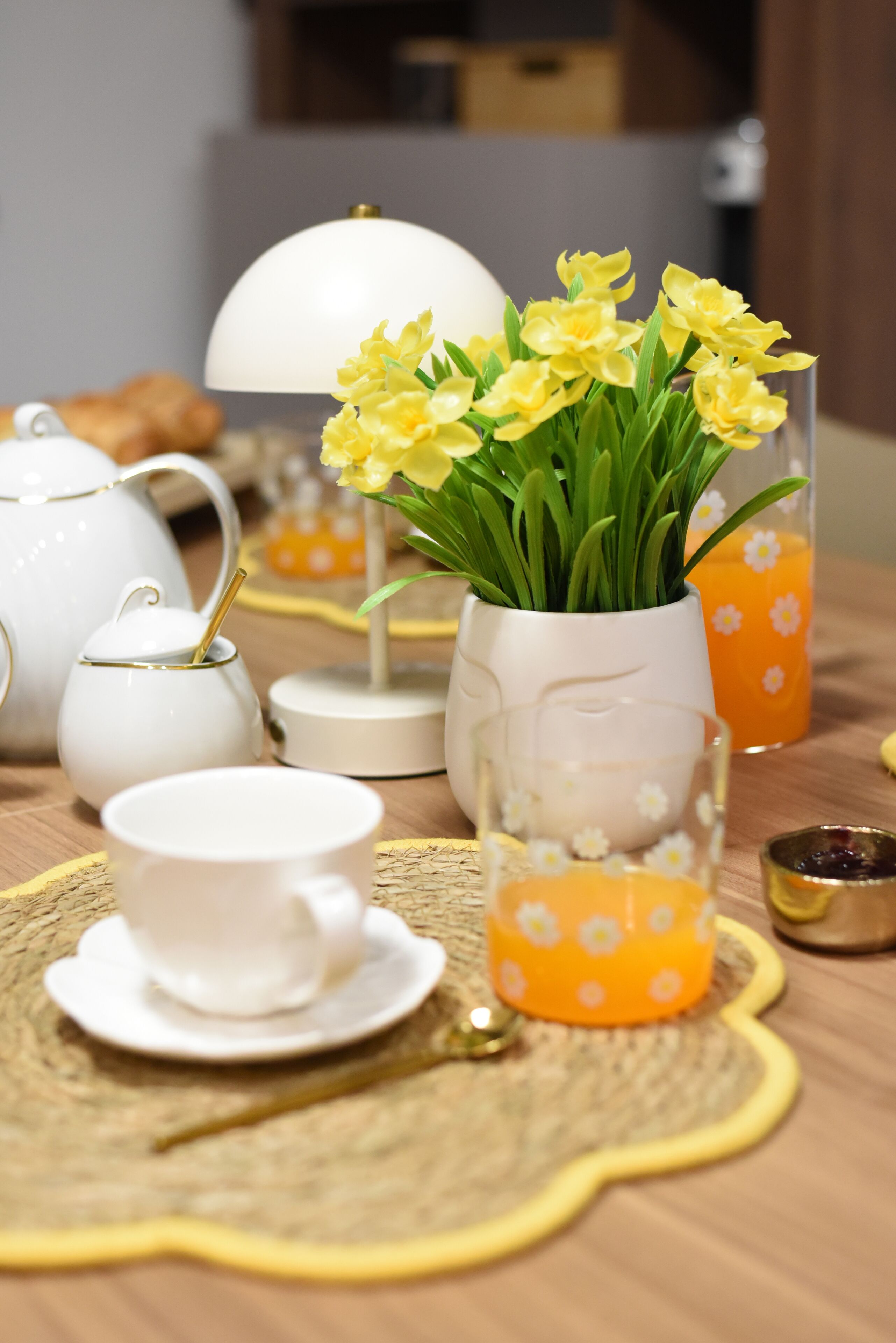 A table set with white teapots, cups, a vase with yellow flowers, and a glass with orange juice.