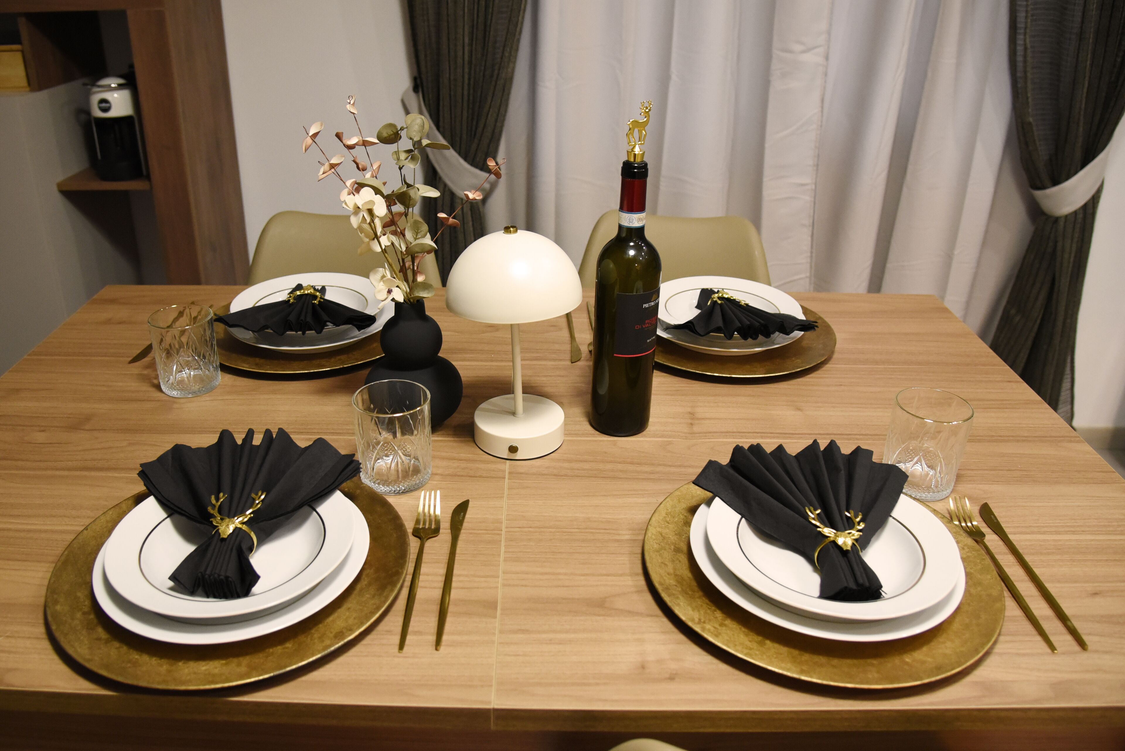 Een eettafel gedekt met witte borden, gouden onderborden, zwarte servetten en een fles wijn.
