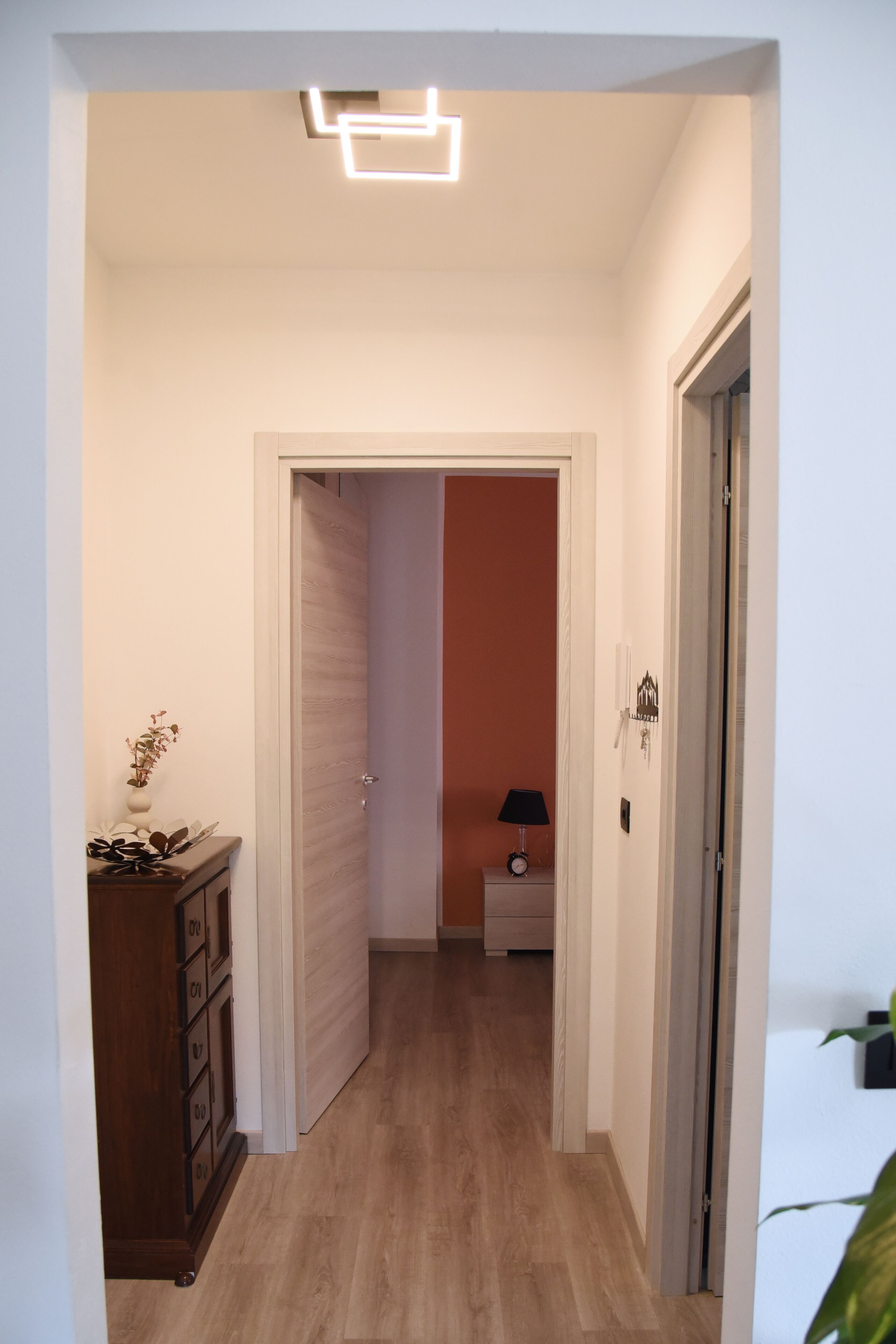 A hallway with a wooden floor, a door with a light switch, and a small table with a lamp.