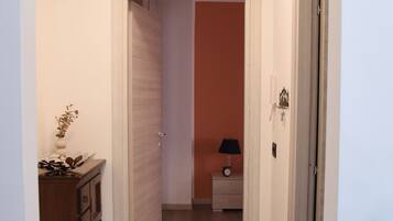 A hallway with a wooden floor, a door with a light switch, and a small table with a lamp.