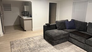 Ein modernes Wohnzimmer mit einem grauen Ecksofa, einem kleinen Couchtisch und einem grauen Teppich. Im Hintergrund ist eine Küchenzeile mit Spüle und Kühlschrank zu sehen.