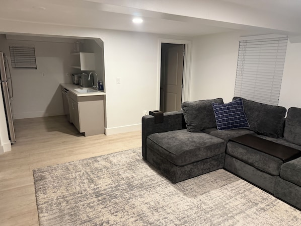 Ein modernes Wohnzimmer mit einem grauen Ecksofa, einem kleinen Couchtisch und einem grauen Teppich. Im Hintergrund ist eine Küchenzeile mit Spüle und Kühlschrank zu sehen.