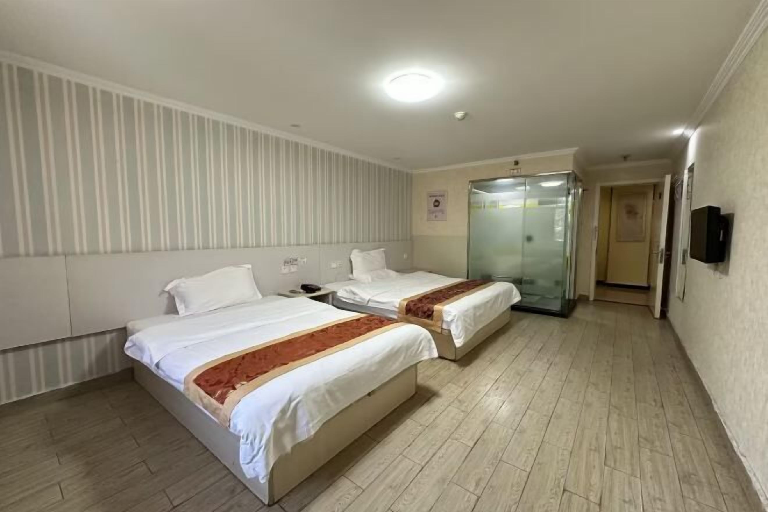 Une chambre d’hôtel avec deux lits, une salle de bain avec une porte en verre et un téléviseur fixé au mur.