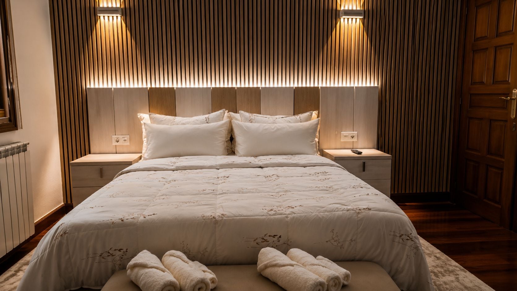 Una cama bien hecha con una colcha floral y dos almohadas, flanqueada por mesitas de noche con lámparas, en una habitación con paneles de madera y una puerta de madera.