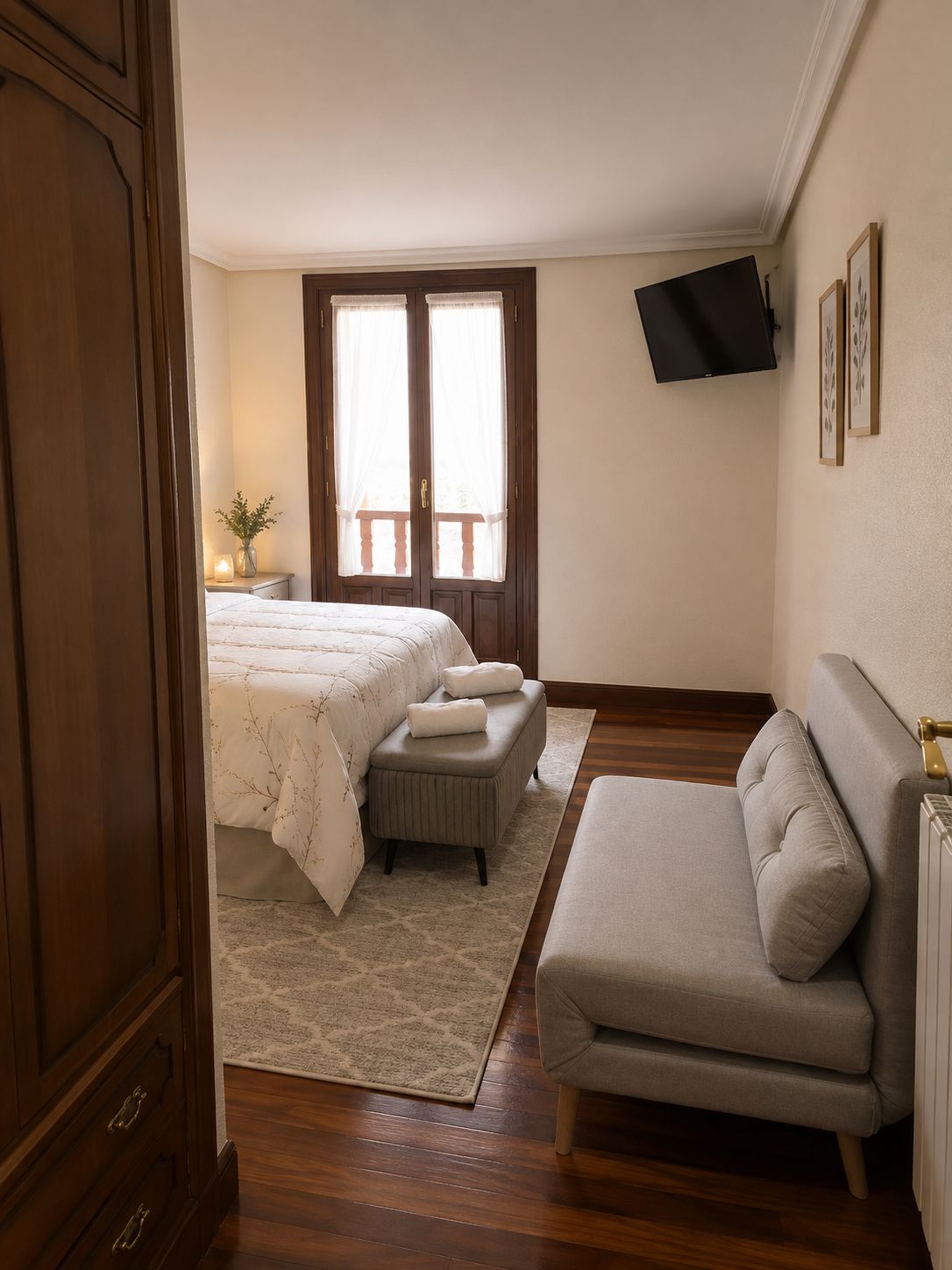 A bedroom with a bed, a sofa, a TV mounted on the wall, and a window with curtains.