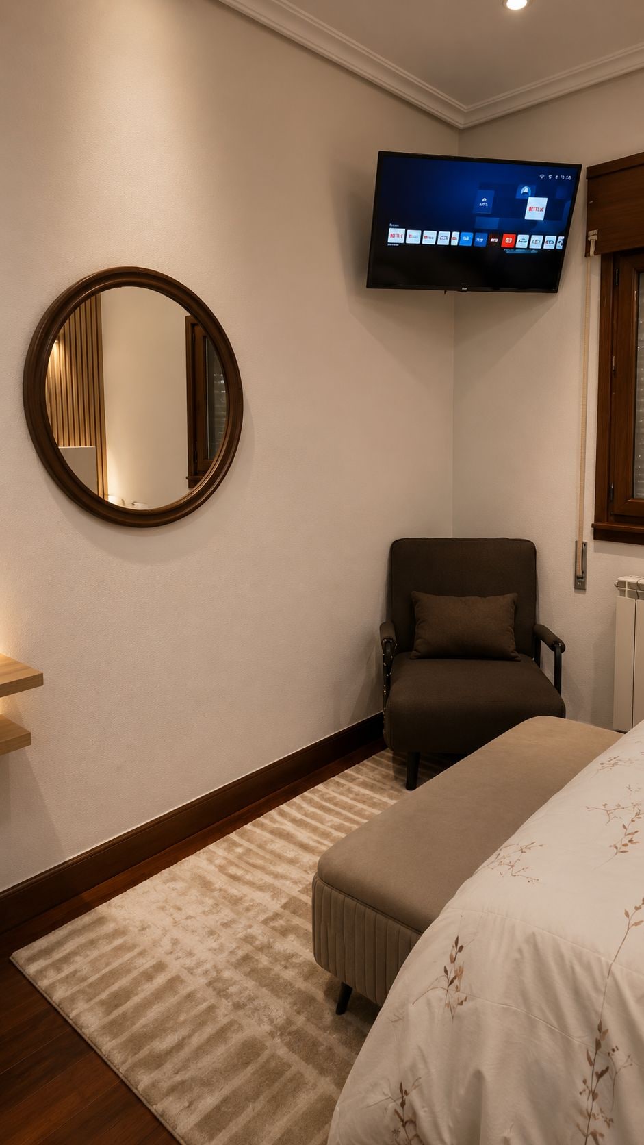 Una habitación de hotel con una cama, una silla, un televisor montado en la pared y un espejo redondo.