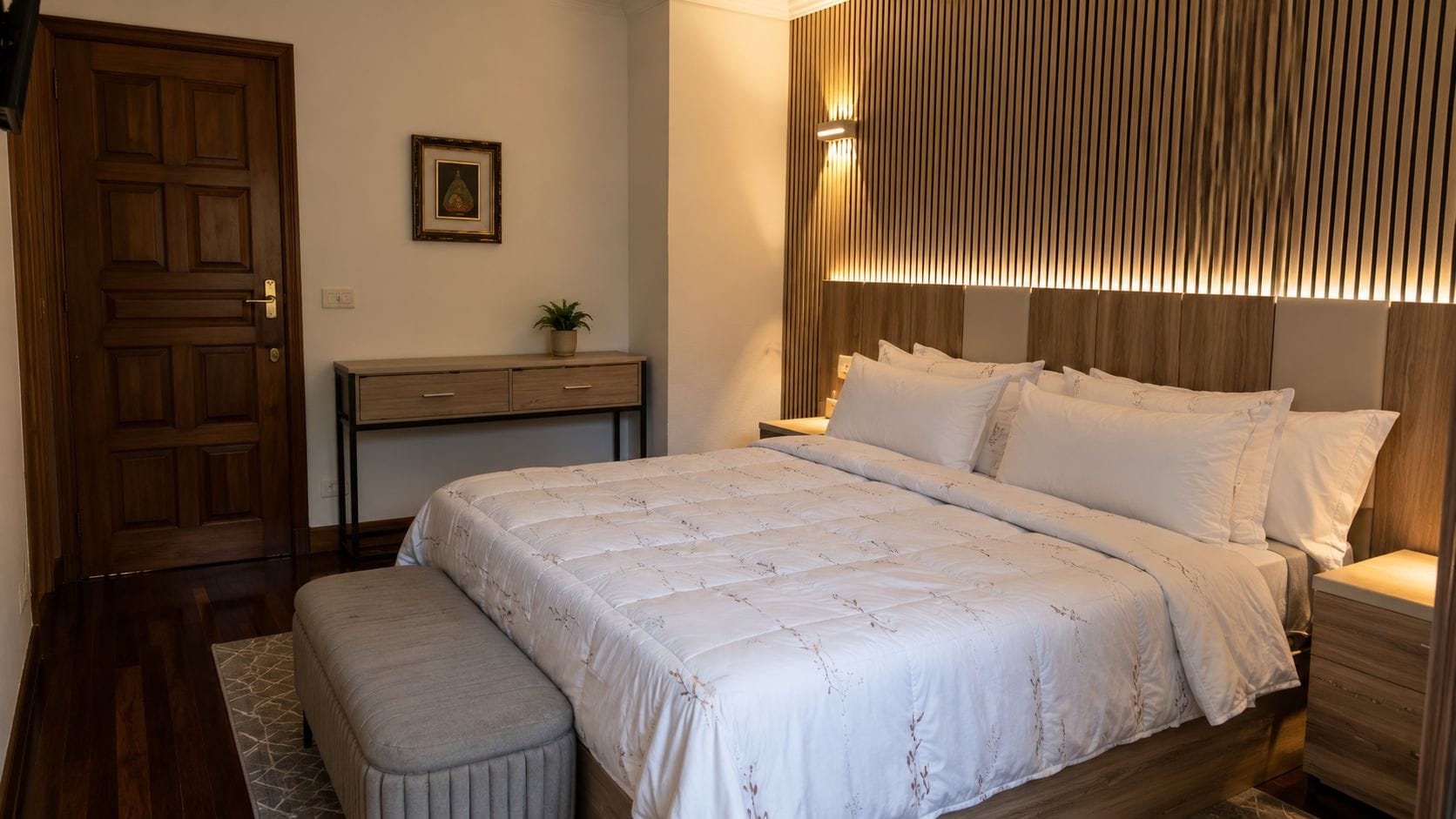 A bedroom with a bed, bedside tables, a nightstand, a wooden door, a small desk with drawers, and a framed picture on the wall.