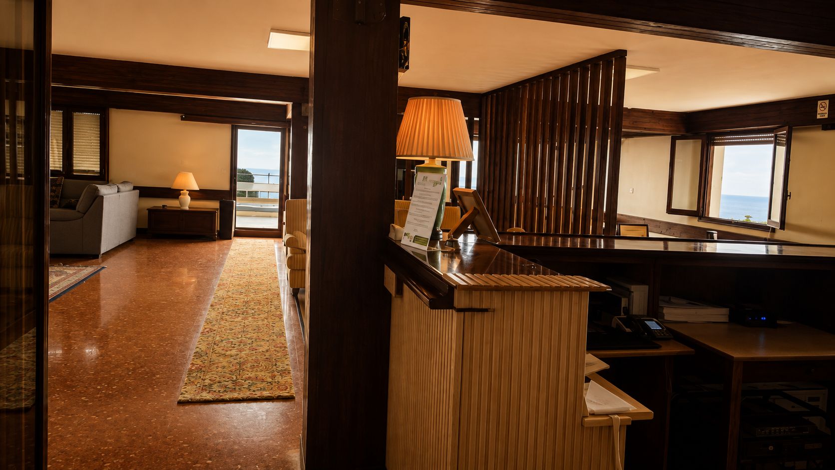Vestíbulo de hotel con recepción, zona de estar con sofá y sillones, pared de paneles de madera y vistas a través de un ventanal grande.