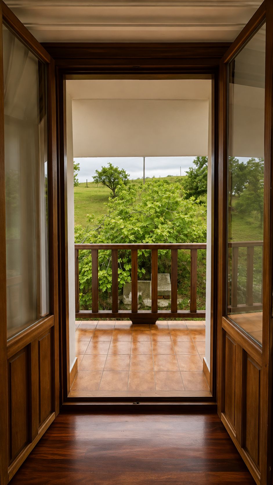 A room with a wooden floor, a balcony with a wooden railing, and a view of a green landscape.