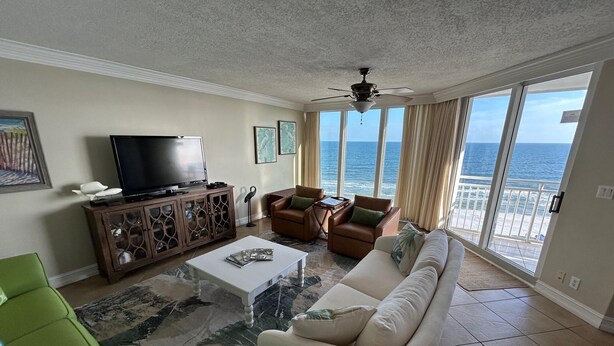 A living room with a television, sofa, and chairs, a ceiling fan, and a view of the ocean.