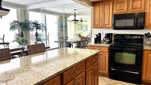 A kitchen with wooden cabinets, a granite countertop, a black microwave, a stove, and a dining area with wicker chairs and a table.