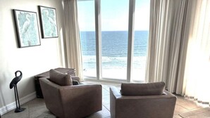 A room with a large window overlooking the ocean, featuring two beige armchairs and a small table.
