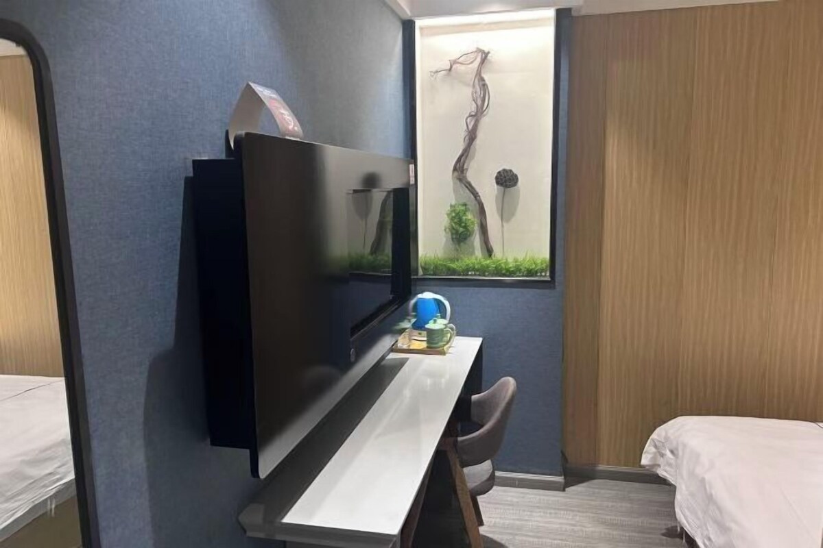 A hotel room with a bed, a desk with a flat-screen TV, a small table with a lamp, and a wall-mounted artwork.