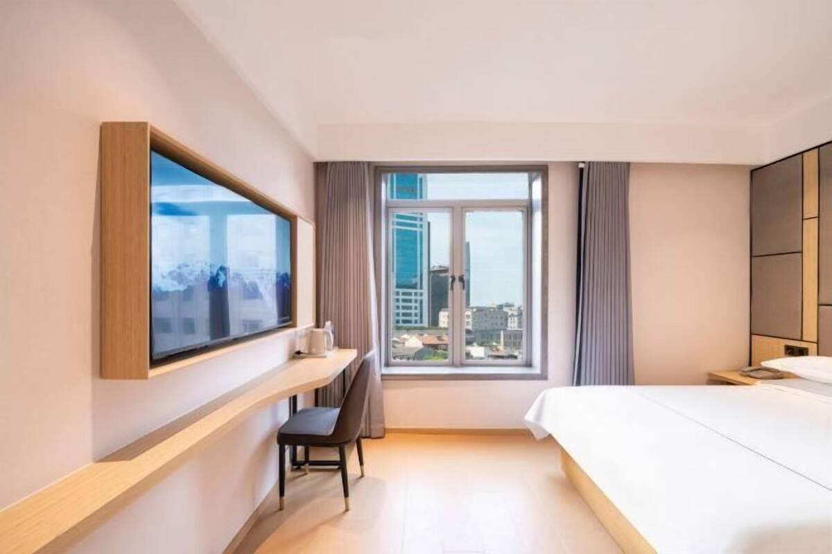 A hotel room with a bed, a desk with a chair, a TV mounted on the wall, and a large window with a city view.