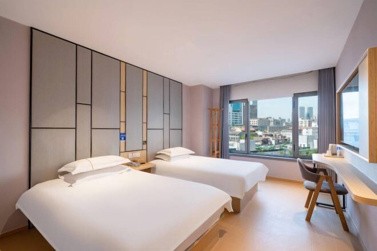 A hotel room with two beds, a desk, and a large window offering a city view.