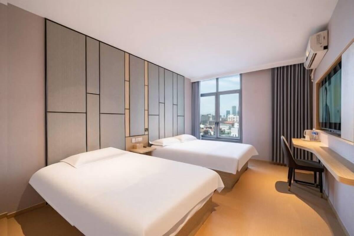 A modern hotel room with two beds, a desk, and a large window offering a city view.