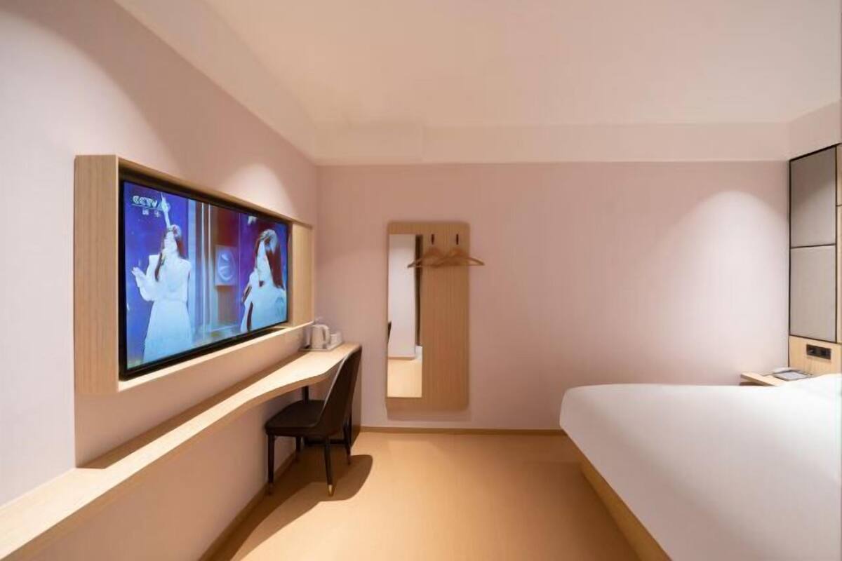 A modern hotel room with a large flat-screen TV, a desk with a chair, a bed, and a wardrobe.