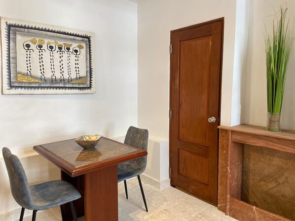 A dining area with a wooden table and two chairs, a framed artwork on the wall, a wooden door, and a plant on a shelf. - Paseo Colonial - Sanctuary 2 (Santo Domingo)