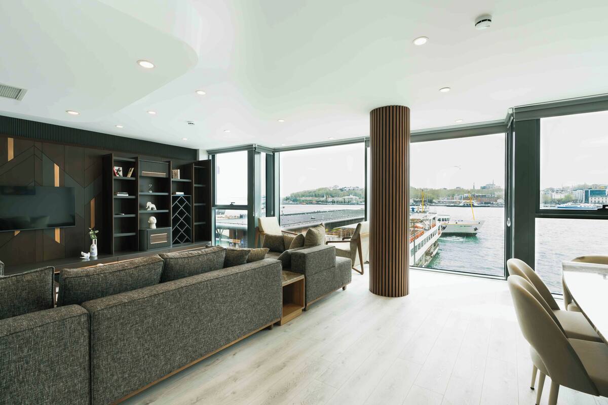 Una sala de estar moderna con un sofá grande, estanterías empotradas, un televisor de pantalla plana y vistas a un río con barcos.