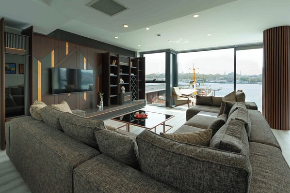 A modern living room with a large sectional sofa, a flat-screen TV, and a view of a cityscape and water through large windows.